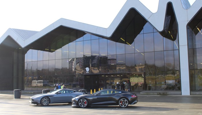 two cars, one grey and one black, parked outside a museum on a car display day