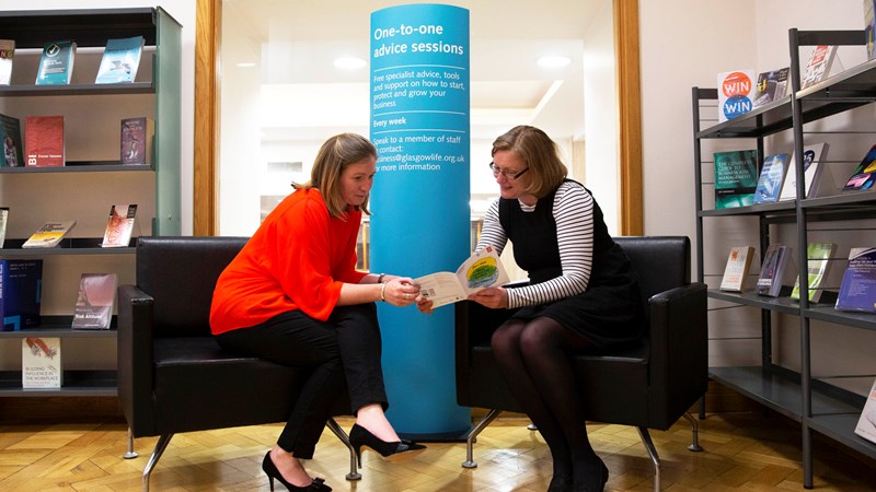 Two women sitting in black armchairs look at a leaflet together.