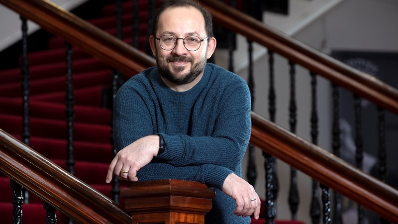 A smiling man leans on the banister of a grand staircase