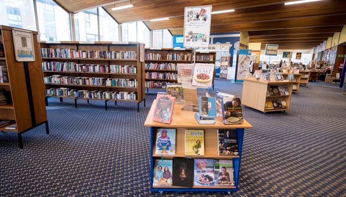 The main library area showing rows of wooden bookshelves, there are standalone bookshelves in the middle of the room. The ceiling is high and has wood pannelled.