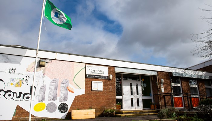 exterior shot of a community centre made of brown brick. there is a sign that says castlemilk community centre in white and black. there is a green flag on a flagpole outside the venue. there is a mural on the side of the build with the work proud printed beside the entrance