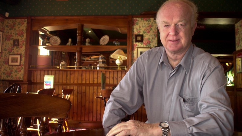 Image of a man called Archie MacPherson sitting at a table looking at the camera in a dimly lit room with a bookcase behind him