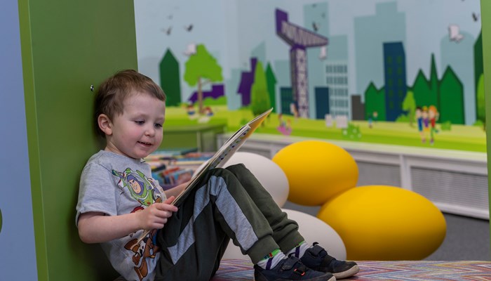 Young child sitting reading a book in Partick library. In the background is a frieze of Glasgow showing a crane, trees and people.