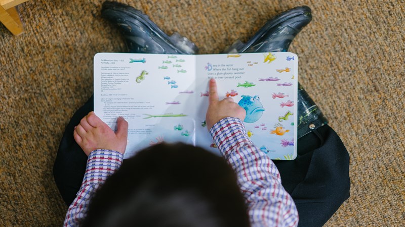 Small child with dark hair, sitting on the floor with their back to the camera reading a book and pointing at the words and fish images. They are wearing wellington boots.