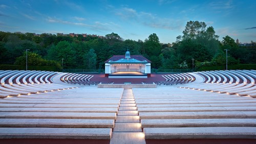 An empty Kelvingrove Bandstand with tall green trees in the background and curved concrete seating, pictured in evening light