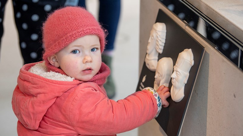 A young child wearing a pink jacket and hat touching a piece of wall art of the wall whilst staring straight into the camera