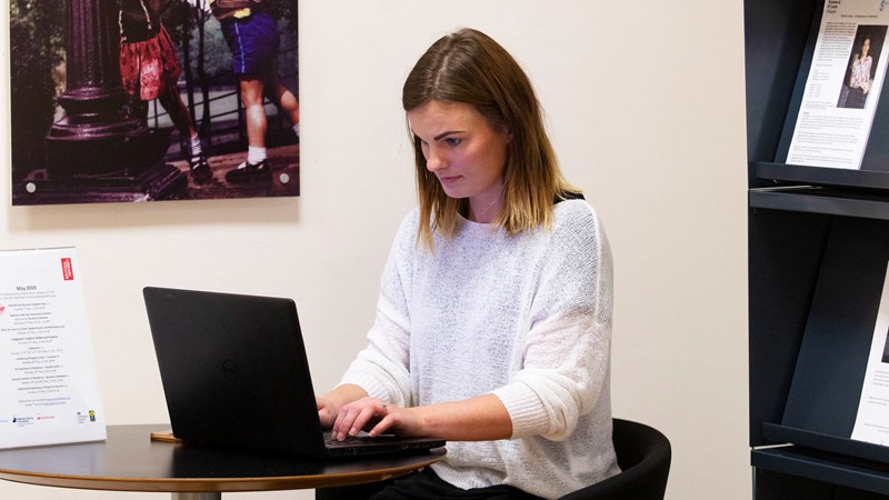 A women sitting at a small desk typing on a laptop with a bookcase behind her with information resources on the shelves