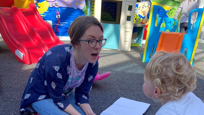 Artist Rebecca Fraser kneels over a home-made banner in a sunny playground, a toddler leaning over to talk to her