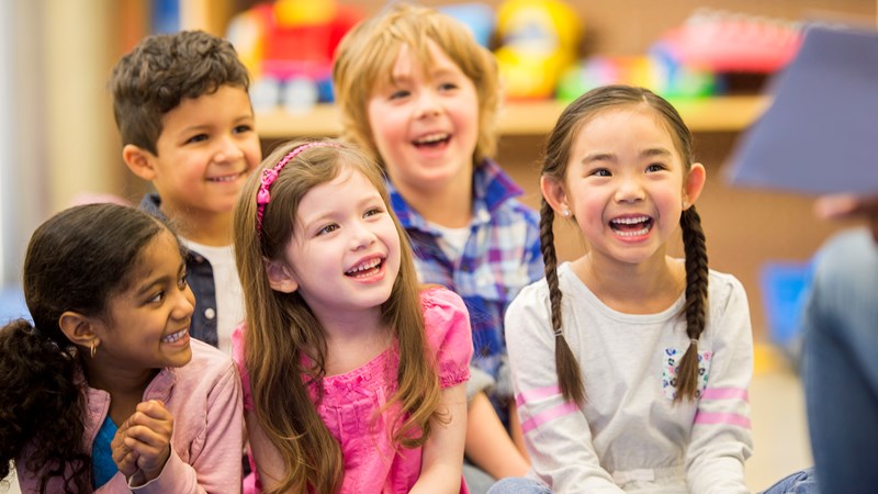 Five seated children all laughing and smiling towards a person (not seen) sitting and reading a book.