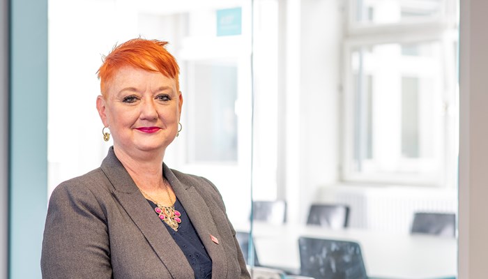 A headshot of Annette Christie, a person with short red hair, wearing a grey tailored jacket, navy top and silver and pink necklace