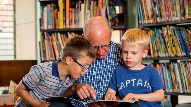 An adult is reading and pointing at a book with a child on each side. They are in a library and there are shelves of books in the background.