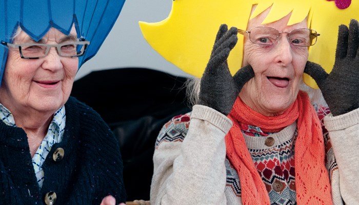 Two people taking part in a workshop. They both have glasses and one has their tongue out and their hands up to their face. The other is moving their hands as if dancing in their seat. One is shown with a big blue hat and the other a yellow type hat with blue. 