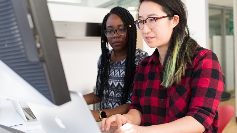 Two people looking at a computer, both with long hair and glasses.