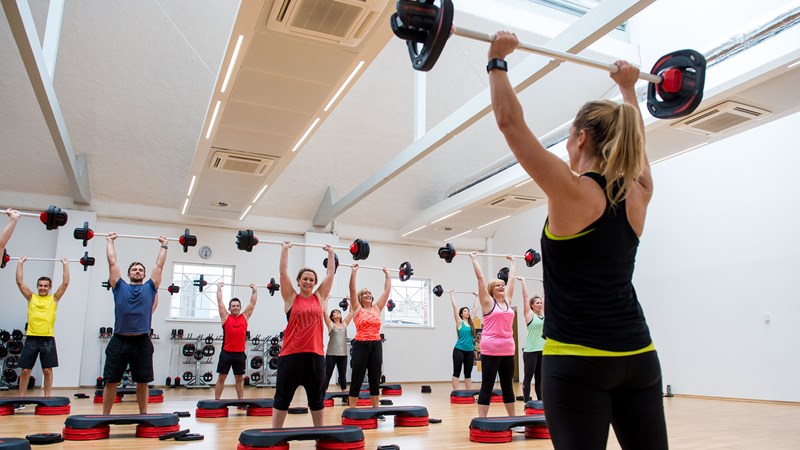 Group of people in a gym class following instructor with barbells above their heads