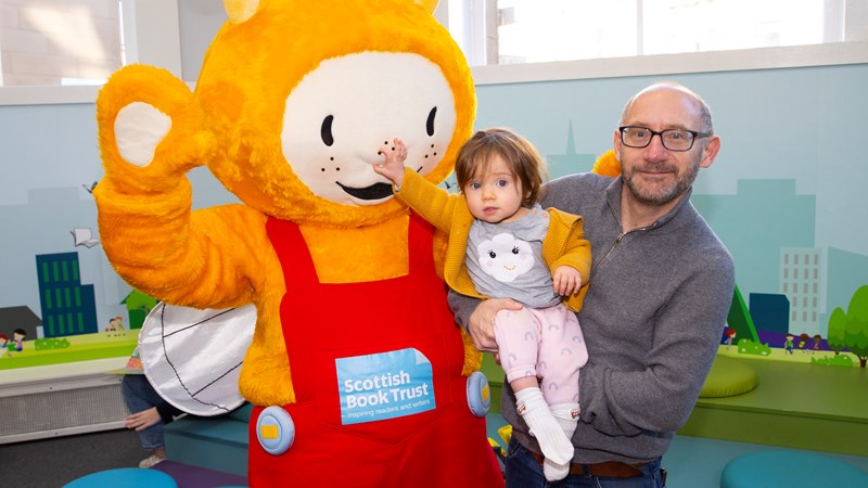 Bookbug in Partick Library. Adult holding a small child who is squeezing the Bookbug mascot's nose. Bookbug is a yellow smiling bug developed by the Scottish Book Trust. They have wings and antennae and wear red dungarees. 