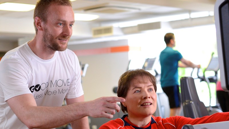 one man standing pointing at a piece of exercise equipment while a women in a wheelchair beside him looks at what he is pointing to