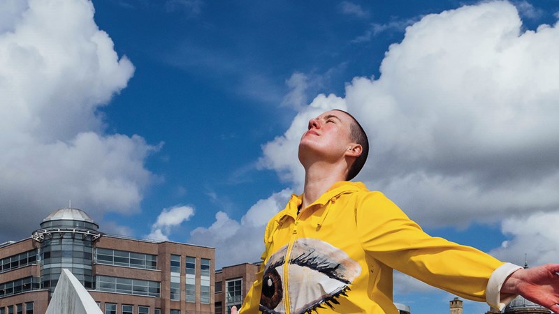 A performer dressed in a yellow jacket with a large eye on the front. They have their face to the sky and arms outstretched behind them. There are other similarly dressed performers in the background. The performance is taken place outdoors on a bridge in Glasgow and there is a blue sky and buildings in the background.