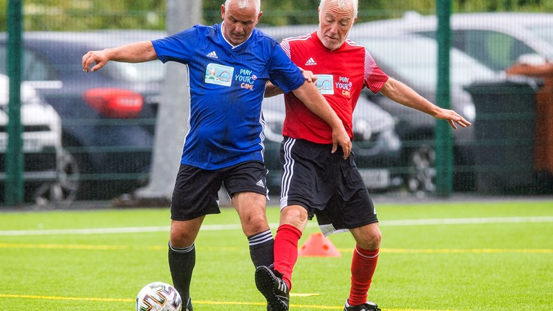 two men tackling each other on an outdoor football pitch with a carpark in the background, one man has the ball at his feet