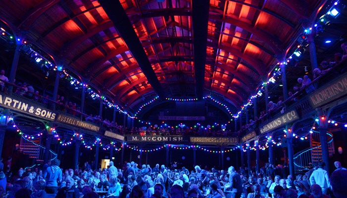 a gig setting indoors with blue lighting throughout. there are lots of people are fairy lights hanging from the balcony that surrounds the room