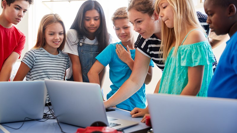 A teacher and a group children gathered round a desk look at a laptop screen, with other laptops on the desk.