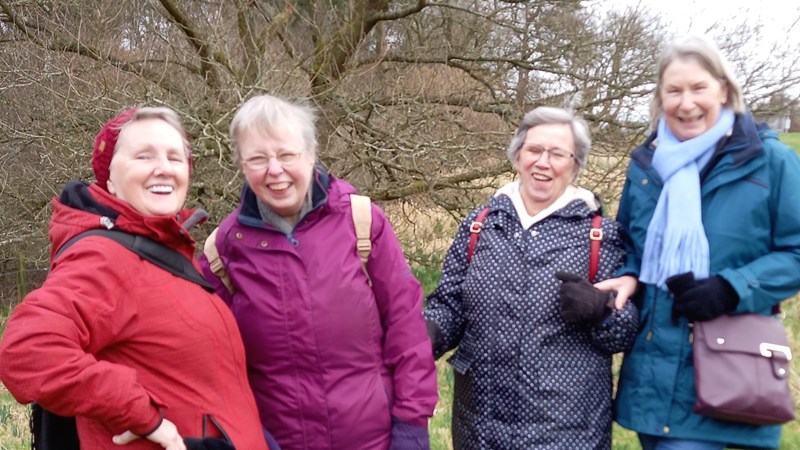 Four people with warm jackets on smiling at the camera . They are standing on grass with bare trees in the background.