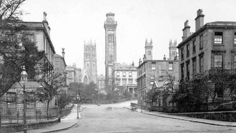 Black and white photograph from early 1900s showing a road, railings, and tall buidlings in the background