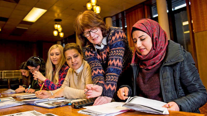 Five women sitting at a desk in a library reading and writing while listening to a tutor who is speaking to them from over their shoulders