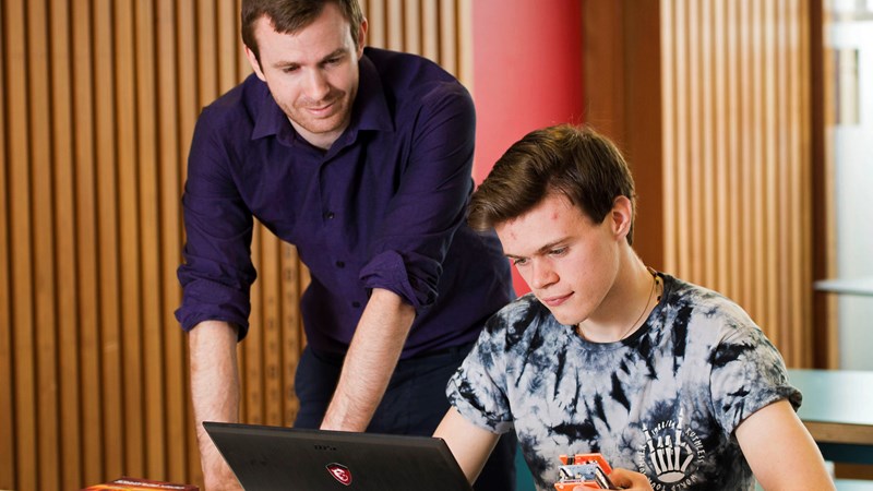 two people, one sitting at a desk with a laptop in front of him and phone in his hand, another person standing looking at the laptop and leaning over. 