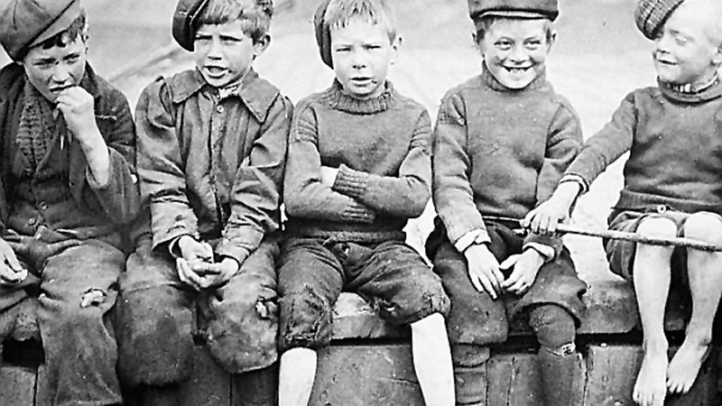 An old black and white photograph of five young people sitting on a wall. They are all wearing flat caps and their clothes are worn. Some do not have shoes on. 