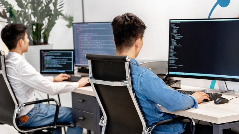 Two students with their backs to the camera sitting in an office chair facing a PC, the screen on the PC showing computer code.